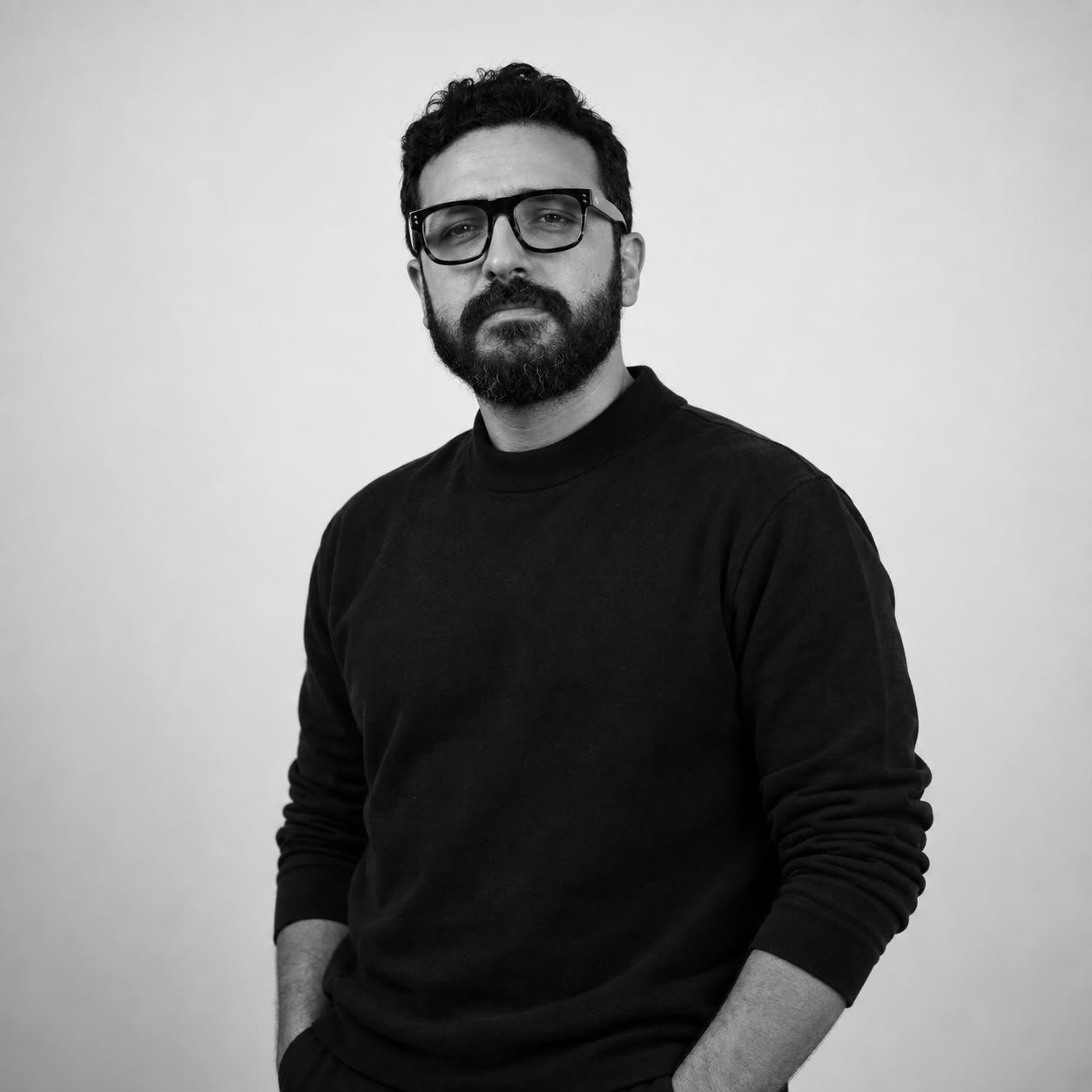 Black-and-white portrait of Eslam Toba in a black crewneck and dark-framed glasses, hands in pockets, against a pale grey studio wall.